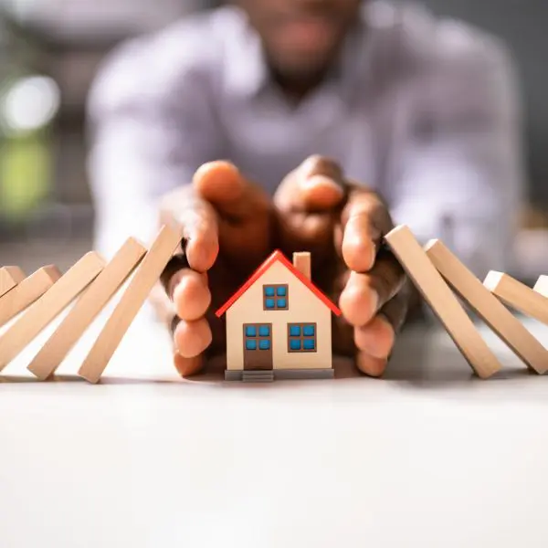 maison miniature en bois avec des dominos en bois qui tombe sur la maison et deux mains qui entourent la maison en signe de protection