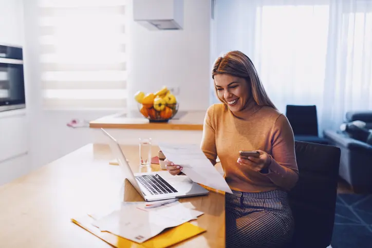 Femme blonde souriante qui regarde un document et dans l'autre mains se trouve son téléphone. Sur la table sont ordinateur portable est posé.