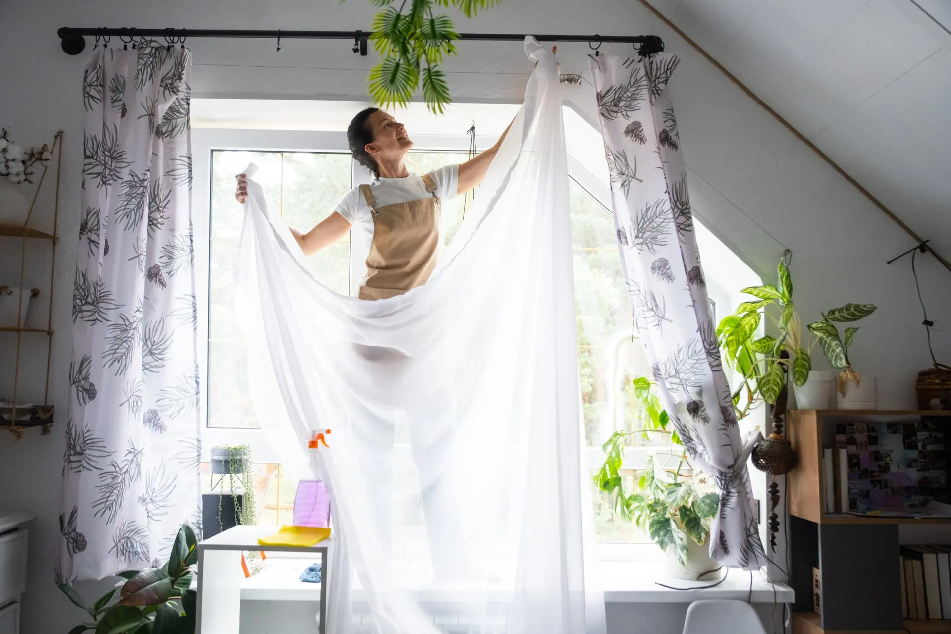La femme en tablier accroche des rideaux de tulle transparent sur de grandes fenêtres mansardées de la maison à l’intérieur de l’intérieur