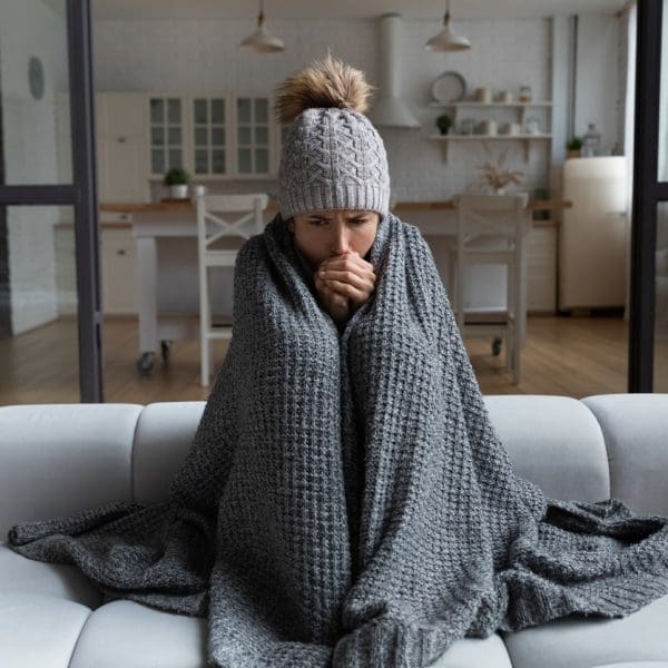 Jeune femme dans une maison, assise sur un canapé avec un bonnet et un plaid sur elle car elle a froid.