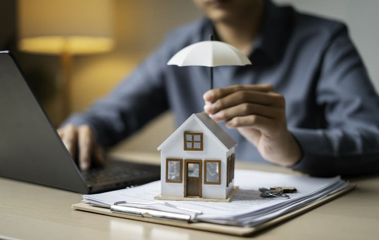 Gros plan d’une petite maison protégée par un parapluie sur des documents de prêt à côté de clés et d’un ordinateur portable, représentant la couverture de la propriété.